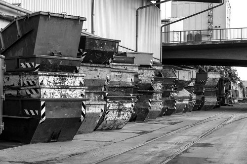 A row of large, industrial metal skips filled with mixed waste materials, positioned on a concrete industrial yard surface adjacent to a warehouse or manufacturing building. The skips are weathered with visible rust and dirt, and are arranged along the side of the building, which features large metal siding and a raised platform with a safety railing in the background. Overhead, there are visible pipes or conduits running along the building's exterior, and the area appears to be part of a commercial waste collection or disposal site in Walthamstow. The ground shows signs of regular use, with some tire marks and minor debris, indicating ongoing waste management activity. The scene is captured in black and white, emphasizing the industrial context and the utilitarian appearance of the rubbish storage containers managed by Commercial Waste Walthamstow, showcasing the typical environment for commercial rubbish collection services in the local area.