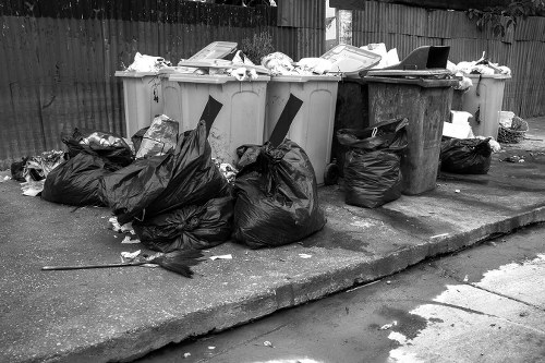 A row of four large wheelie bins positioned on a concrete pavement, with two grey and one brown bin, all filled with assorted rubbish. In front of the bins, numerous black plastic rubbish bags are piled up, some somewhat torn or bulging, indicating full contents. The surface around the bins shows signs of use, with scattered debris and discarded paper. In the background, a wooden fence runs horizontally, providing a boundary for the area. The scene appears to be outdoors, likely in a residential or commercial setting within Walthamstow, suggesting a waste collection or rubbish removal activity by Commercial Waste Walthamstow. The lighting is natural, with diffuse daylight illuminating the scene, highlighting the textures of the plastic bags, bin surfaces, and the concrete pavement. This image illustrates typical waste disposal and rubbish clearance, emphasizing the importance of professional rubbish removal services in Walthamstow to manage household or commercial waste efficiently and tidily.