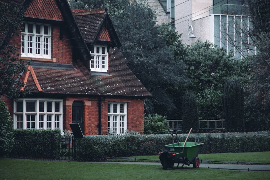 A green wheelbarrow with red handles and wheels is positioned on a paved pathway in front of a red-brick residential house with a sloped, tiled roof and white-framed windows. The wheelbarrow contains 
