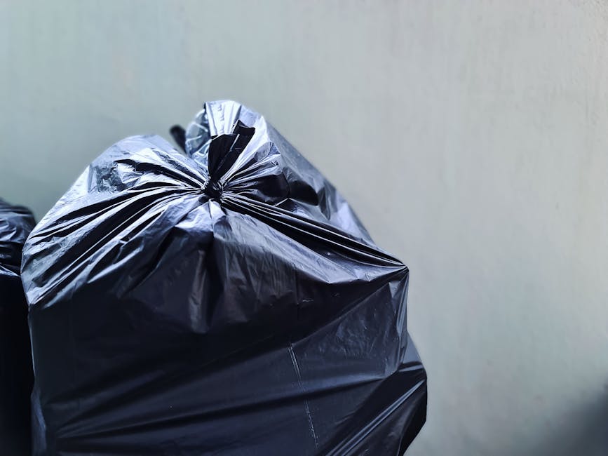 Close-up view of several large black bin bags made of shiny, crinkled plastic material, filled with waste and tightly knotted at the top, resting against a plain, light-colored wall. The bags are plac