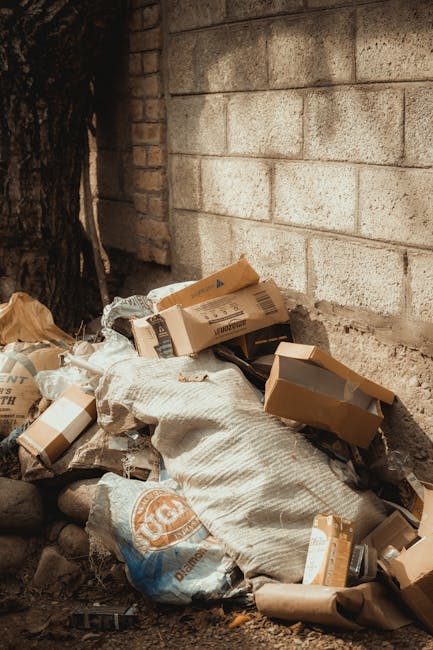 Close-up view of several large black bin bags made of shiny, crinkled plastic material, filled with waste and tightly knotted at the top, resting against a plain, light-colored wall. The bags are plac